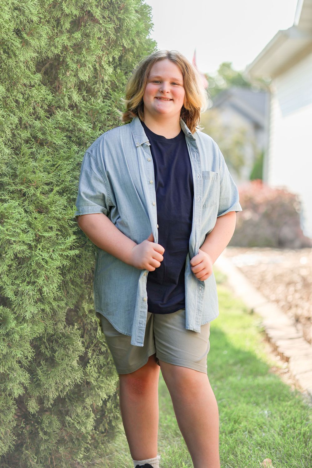 Person in light blue button-down shirt and shorts standing against evergreen bushes outdoors.