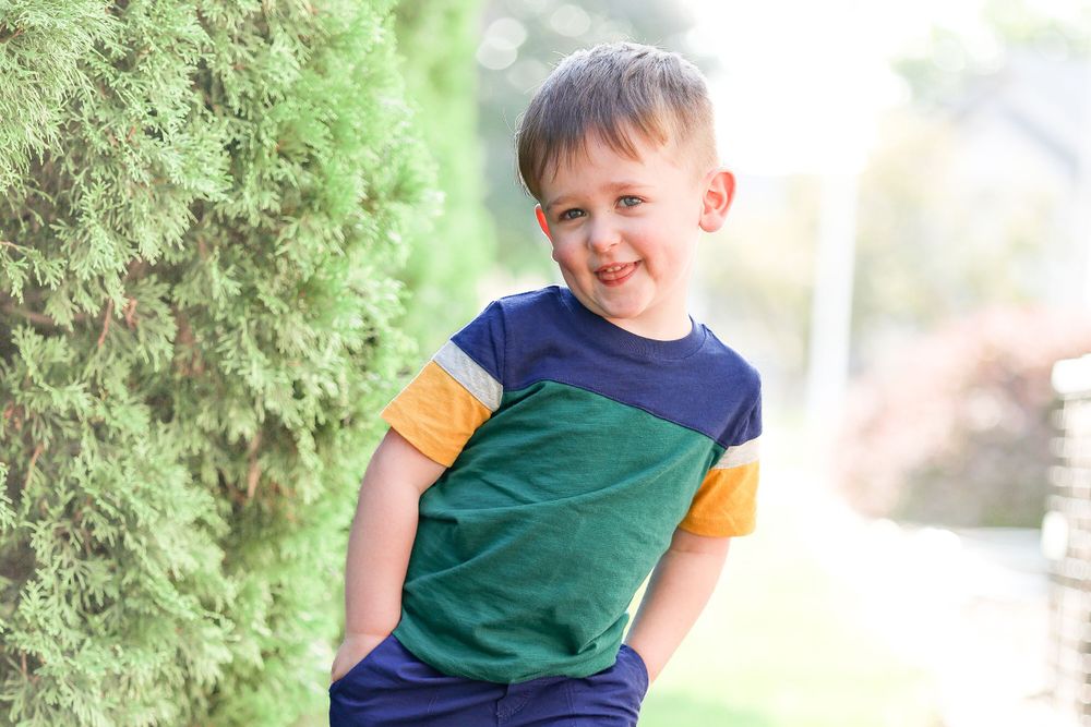 Child in colorblock t-shirt poses confidently against evergreen bushes.