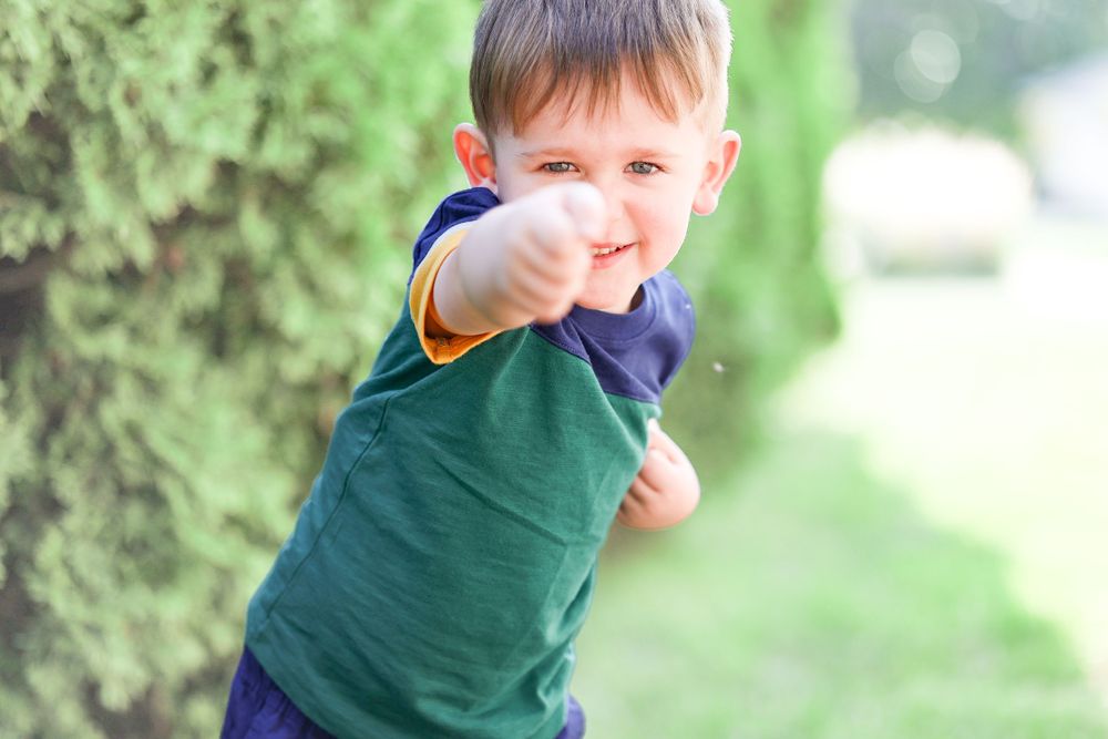 Playful child in navy and green t-shirt giving thumbs up outdoors.