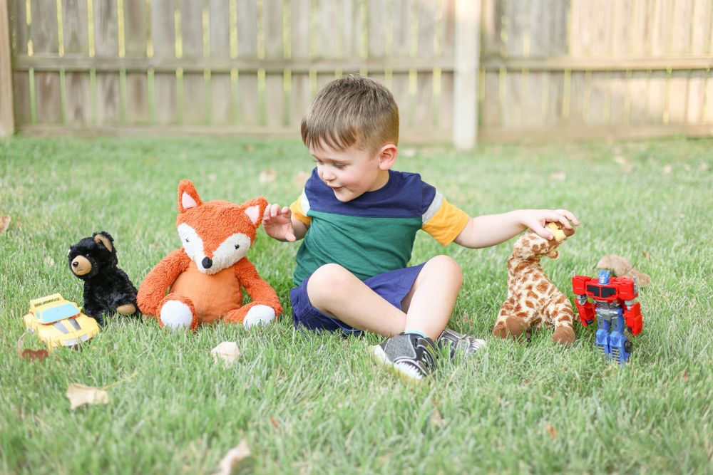 A child sits on grass playing with stuffed animals and toys including a plush fox and giraffe in a backyard.