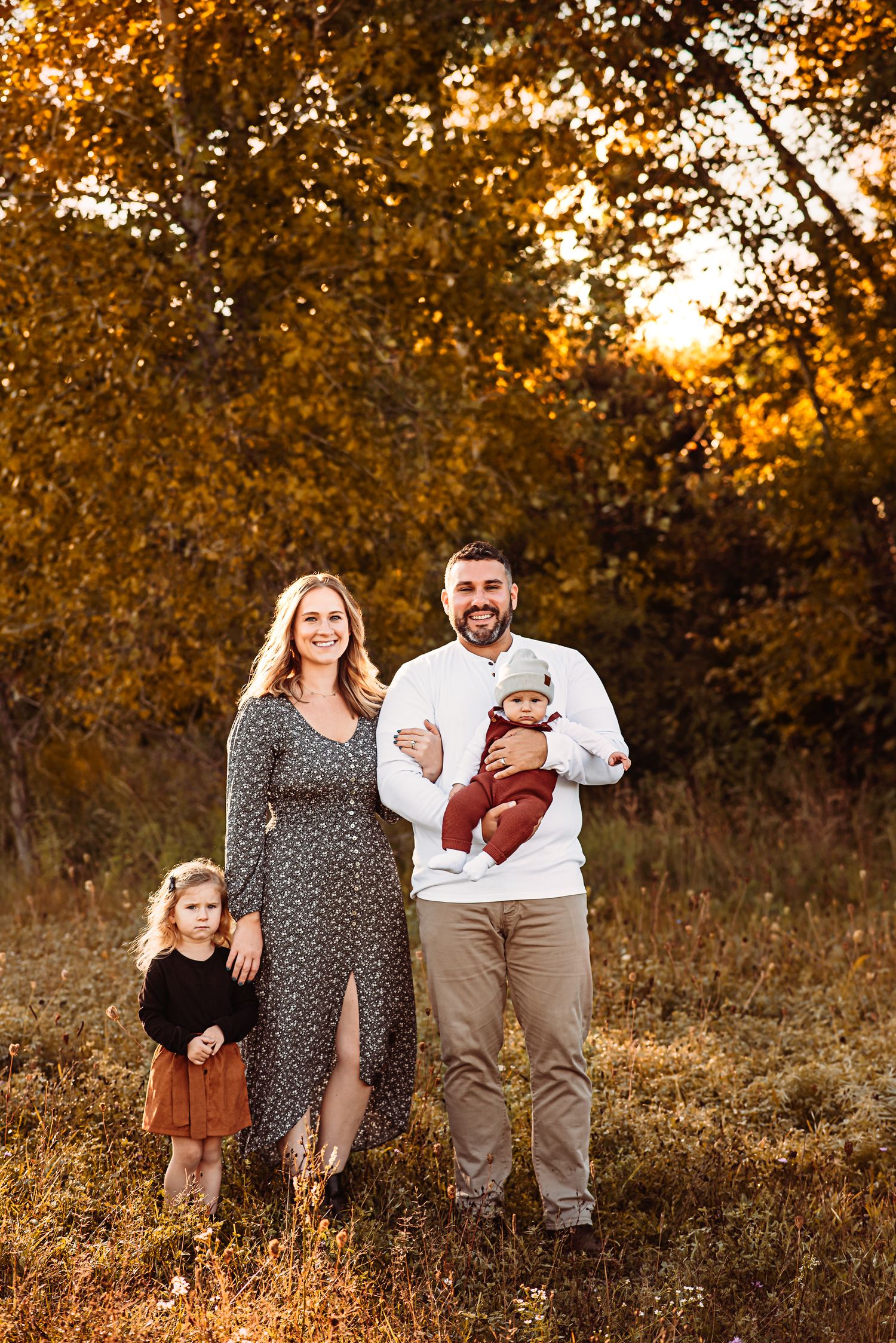 Family portrait at sunset with autumn trees and golden light in background.
