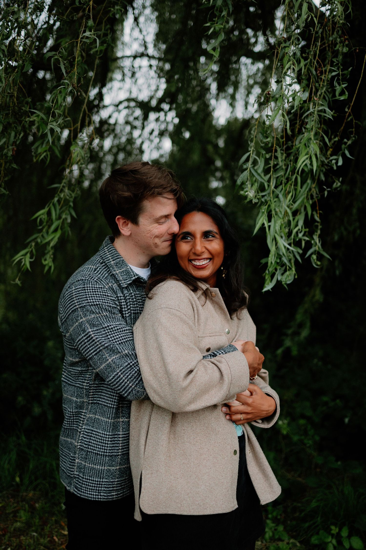 A romantic couple embraces under drooping willow branches in a moody outdoor portrait session.