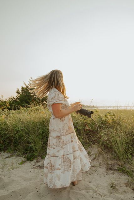 Female photographer in floral dress walking through beach dunes at golden hour.