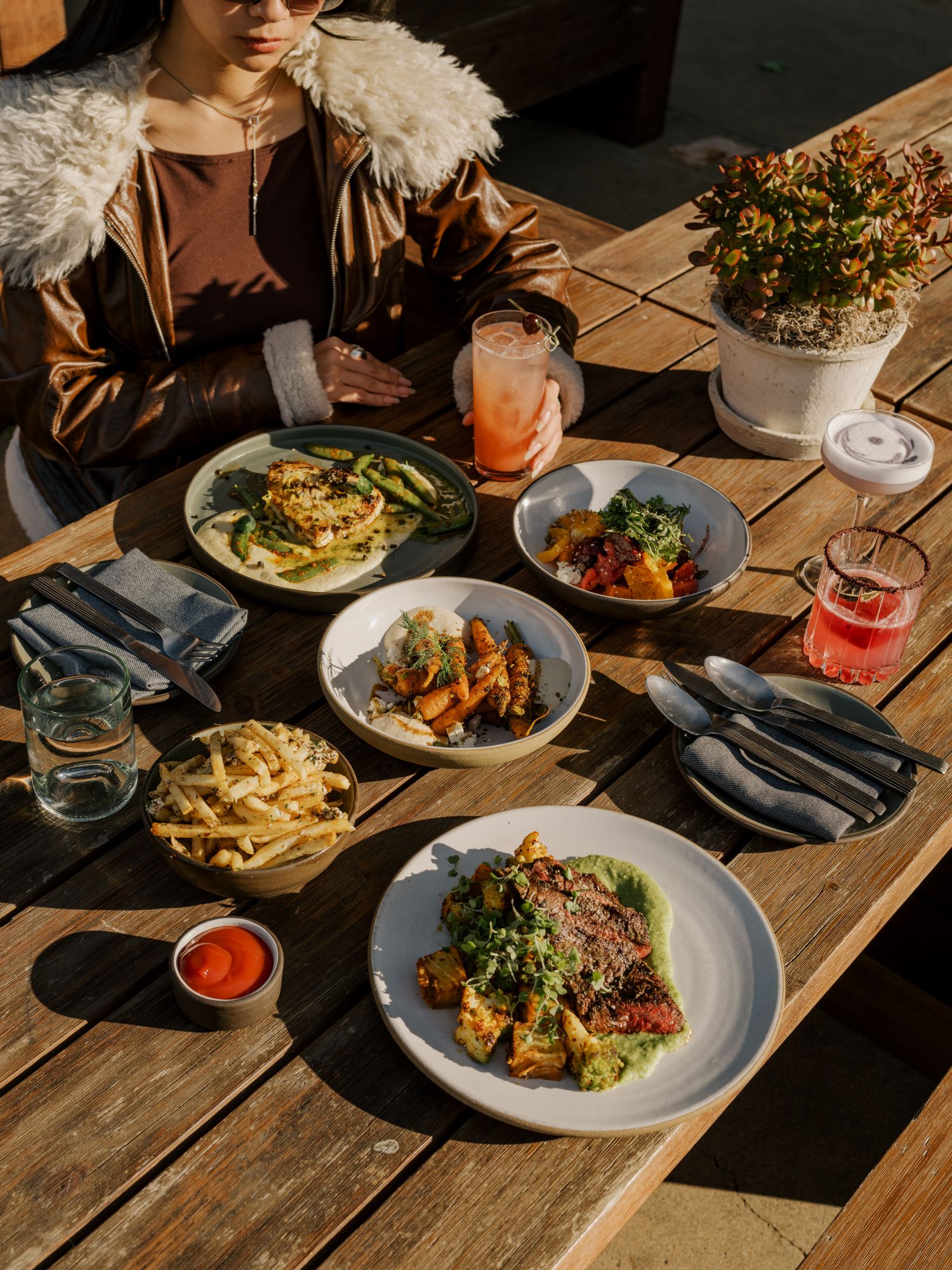 Table spread with various colorful dishes including salads, fries and cocktails in warm sunlight.