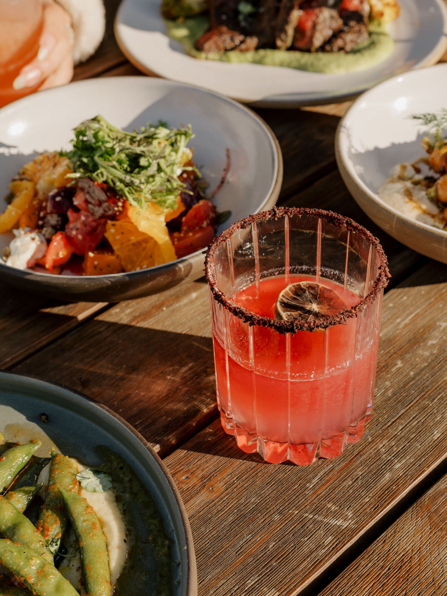 Several gourmet dishes and a vibrant red cocktail with garnish served on a wooden dining table.