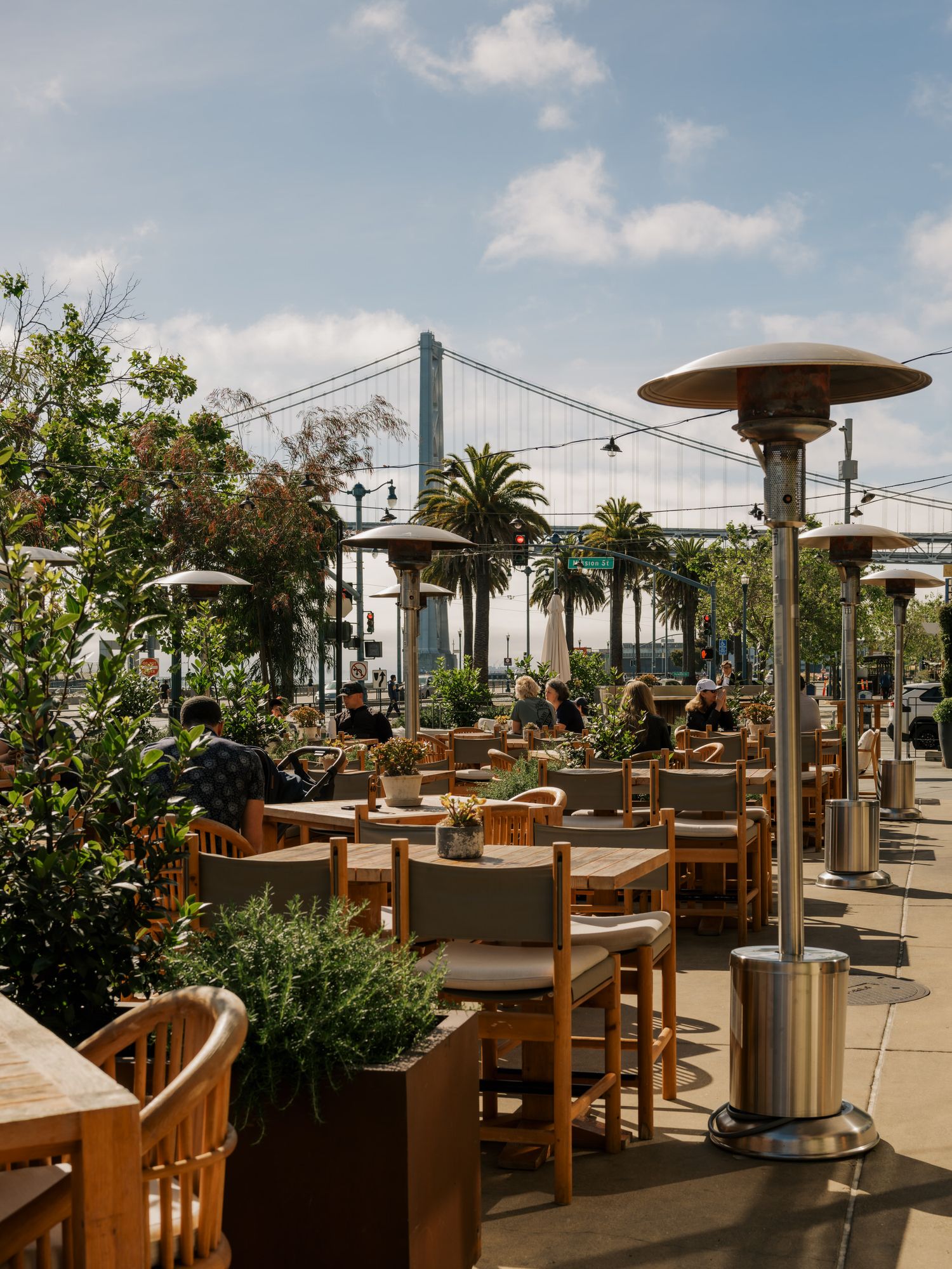 Outdoor restaurant patio with wooden furniture, heat lamps, and palm trees overlooking the San Francisco Bay Bridge.