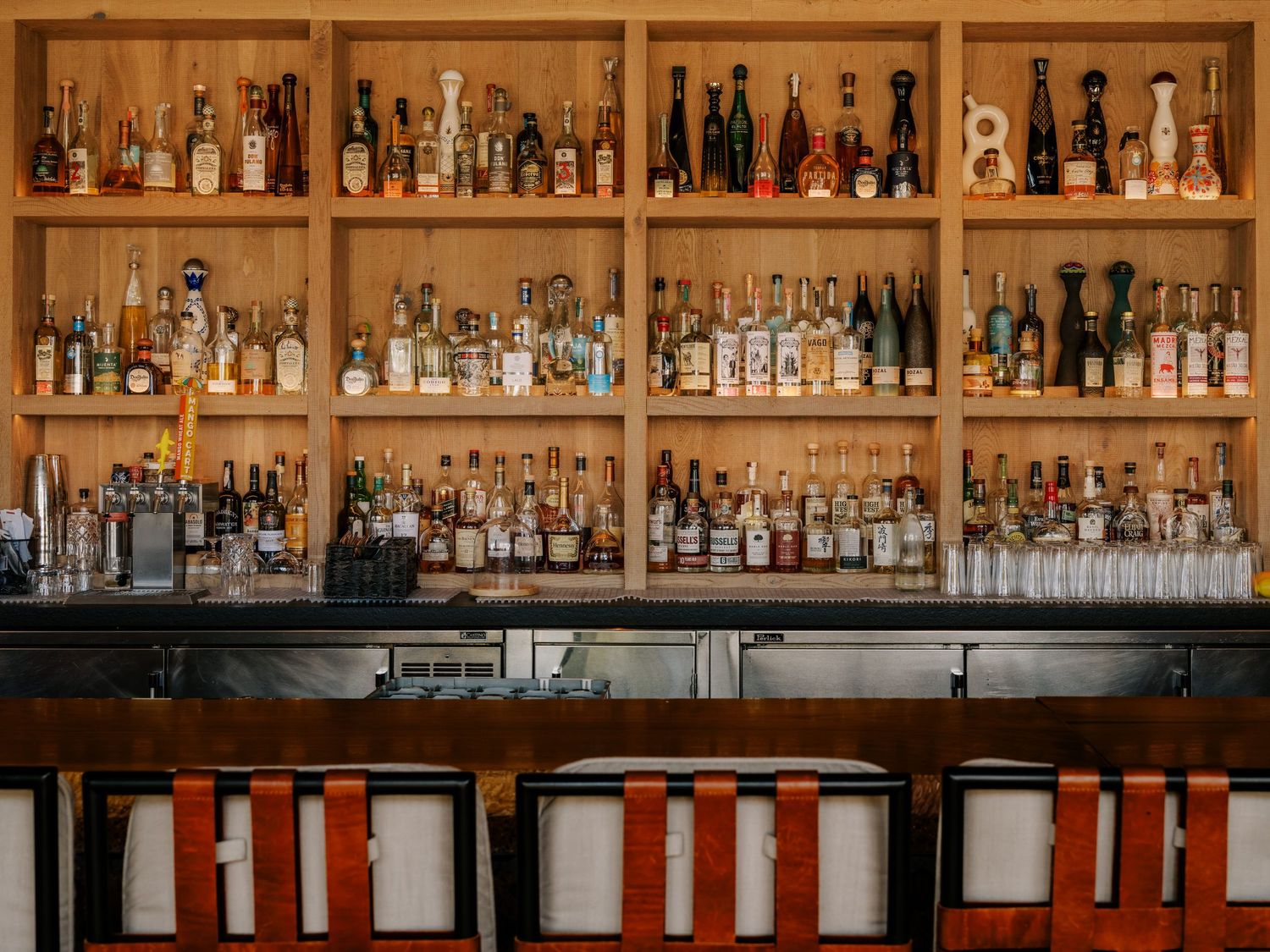 Well-stocked wooden bar shelving displays rows of liquor bottles above red and white industrial bar stools.