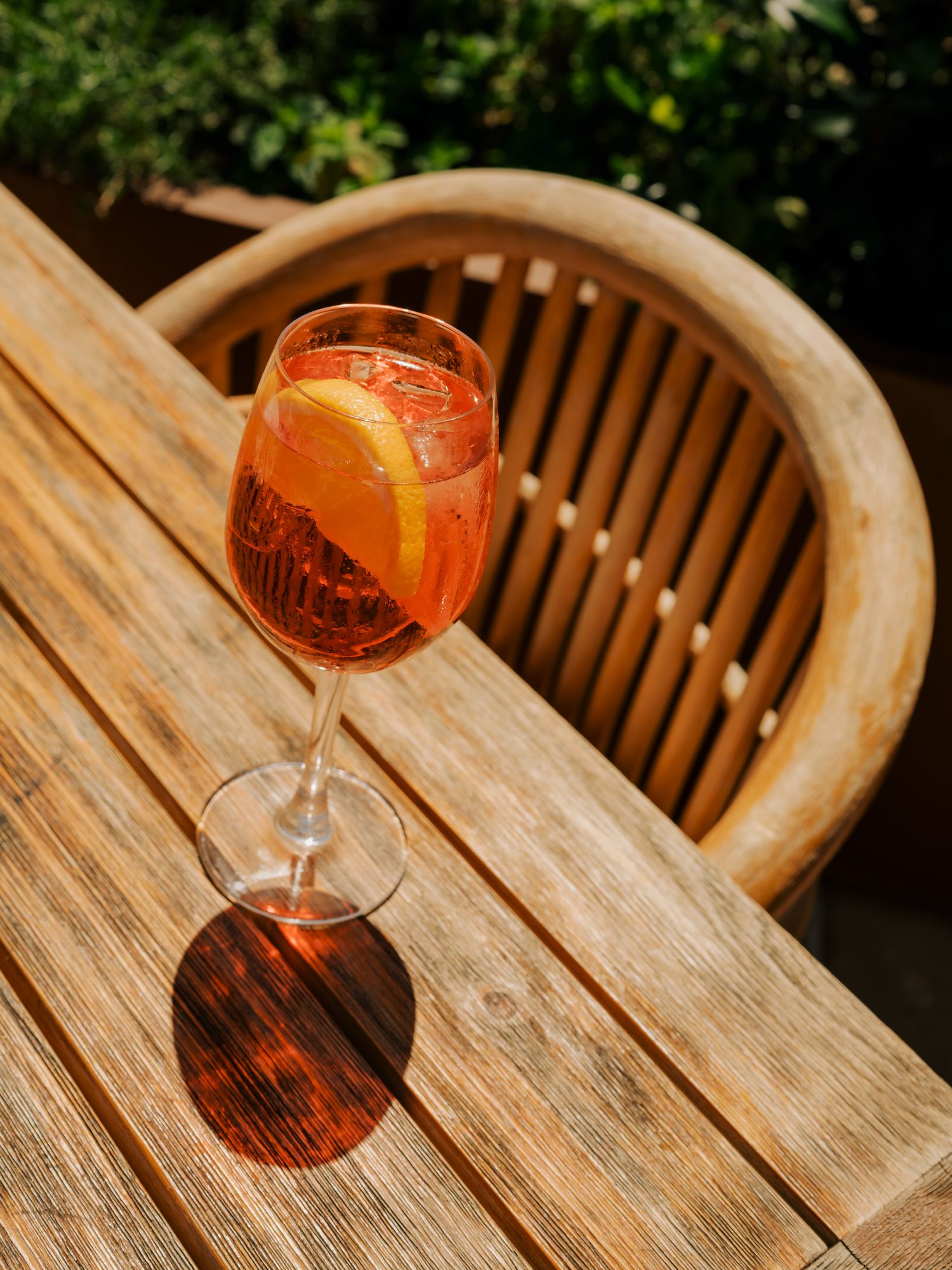 Orange Aperol Spritz cocktail casting a shadow on wooden outdoor table in sunlight.