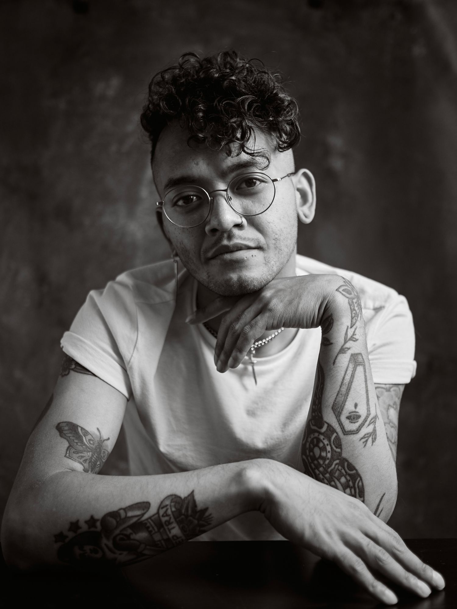 Black and white portrait of a person with curly hair, glasses and tattoos wearing a white t-shirt.