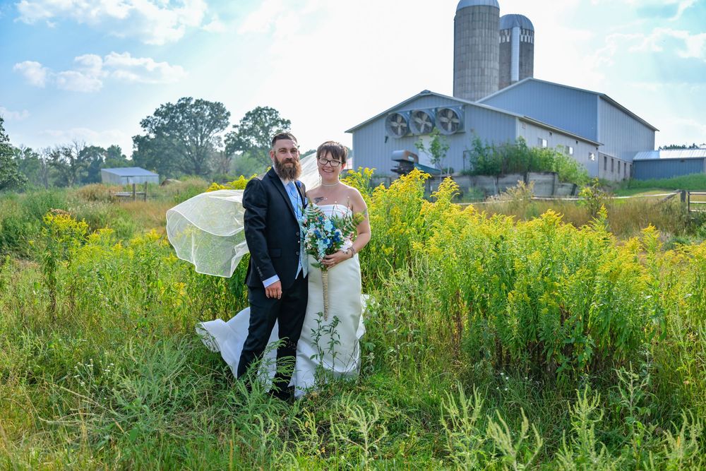 Bride and groom pose in a lush green field with a rustic barn and silos in the background on their wedding day.