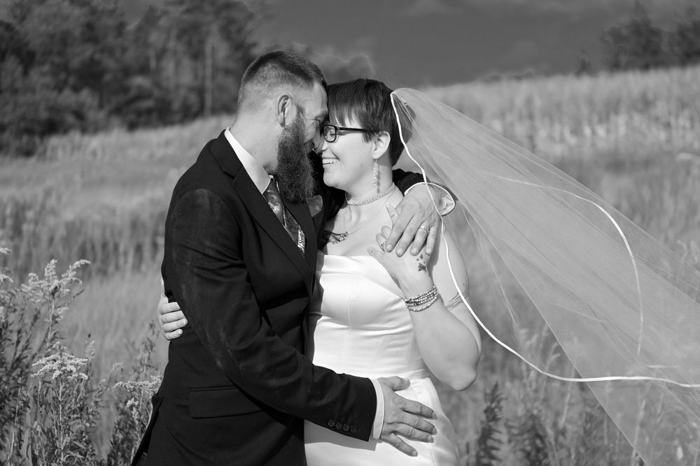 Black and white wedding photo of a couple embracing in a field, bride in veil and groom in suit, smiling at each other.