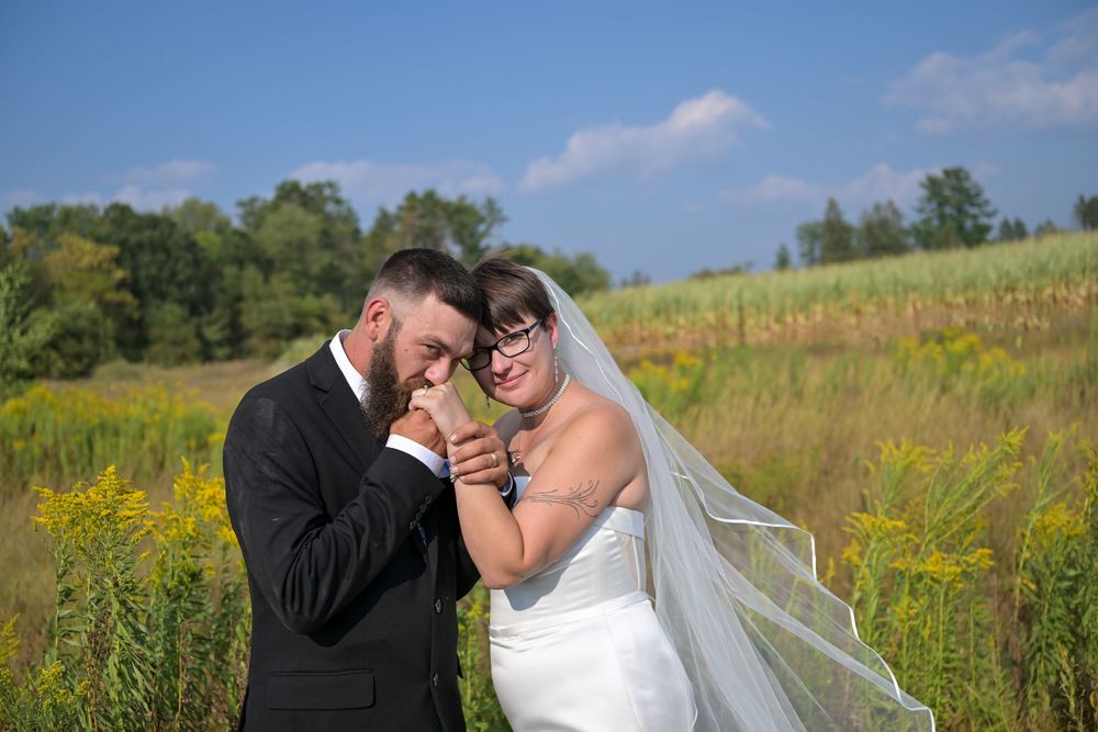 Bride and groom share a tender moment in a sunny field, groom kissing her hands, wearing wedding attire outdoors.