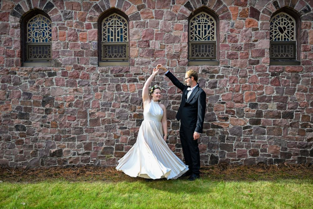 Bride and groom dance joyfully in front of a rustic stone church wall on a sunny day.