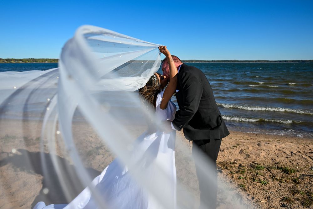 Bride and groom kissing on a beach with veil blowing in the wind against a blue sky and ocean backdrop.
