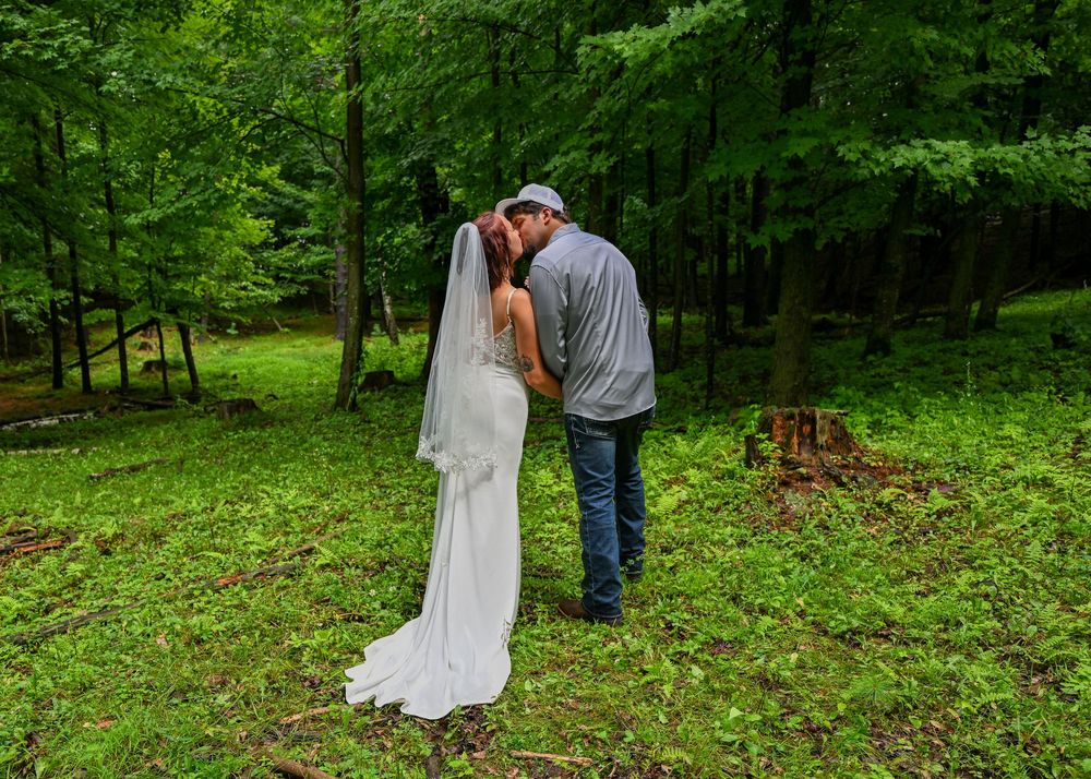 Bride in white gown and veil kissing groom in gray shirt in a beautiful green woodland setting.