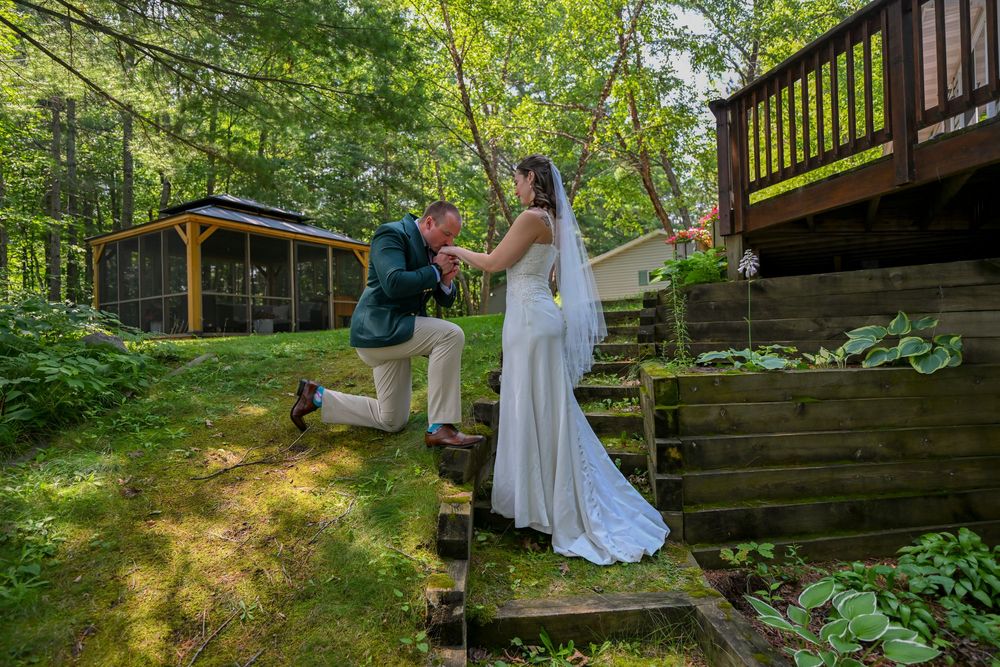 Groom kneels to adjust bride's dress on rustic garden steps beside a wooded cabin on their wedding day.
