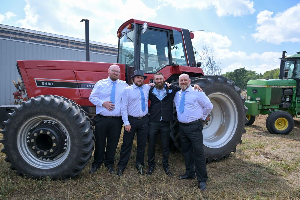 Four men in formal attire pose together in front of a red Case IH tractor at an outdoor farm wedding.