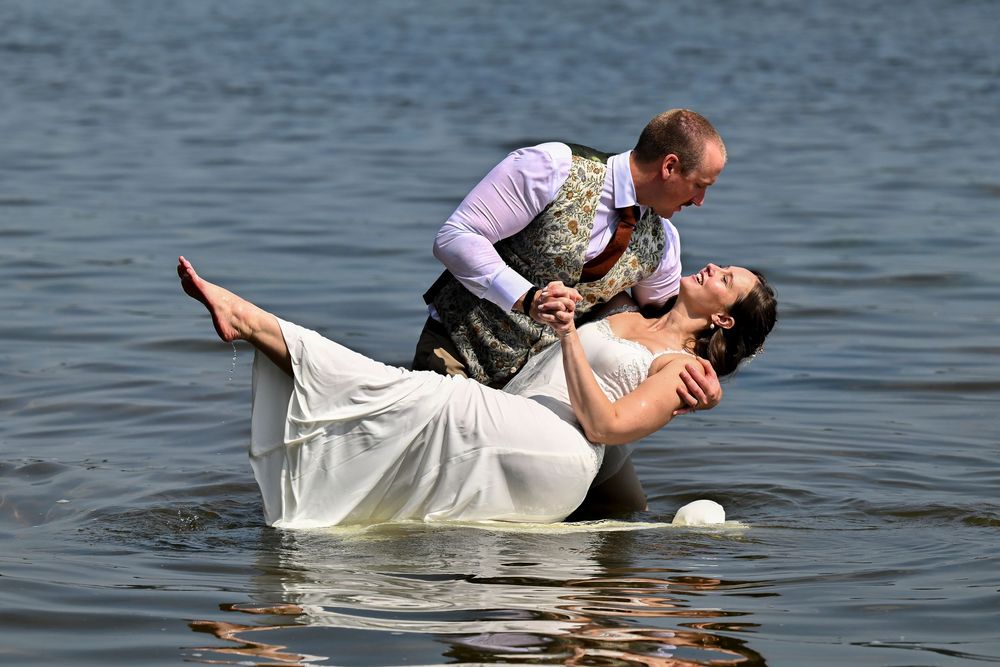 Groom dipping bride dramatically over lake water during a romantic outdoor wedding photo shoot.