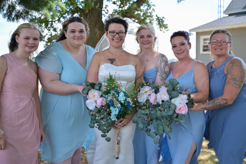 Bride in white strapless gown poses with five bridesmaids in blue and pink dresses holding floral bouquets outdoors.