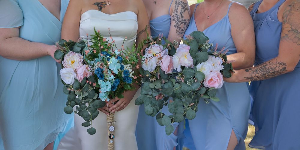 Bride in white gown and bridesmaids in blue dresses holding eucalyptus and pink rose bouquets outdoors.