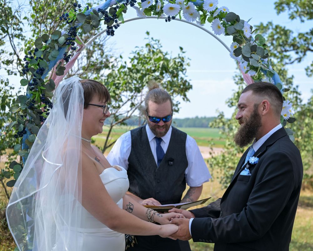 Bride and groom exchange rings outdoors under floral arch with officiant during sunny wedding ceremony.