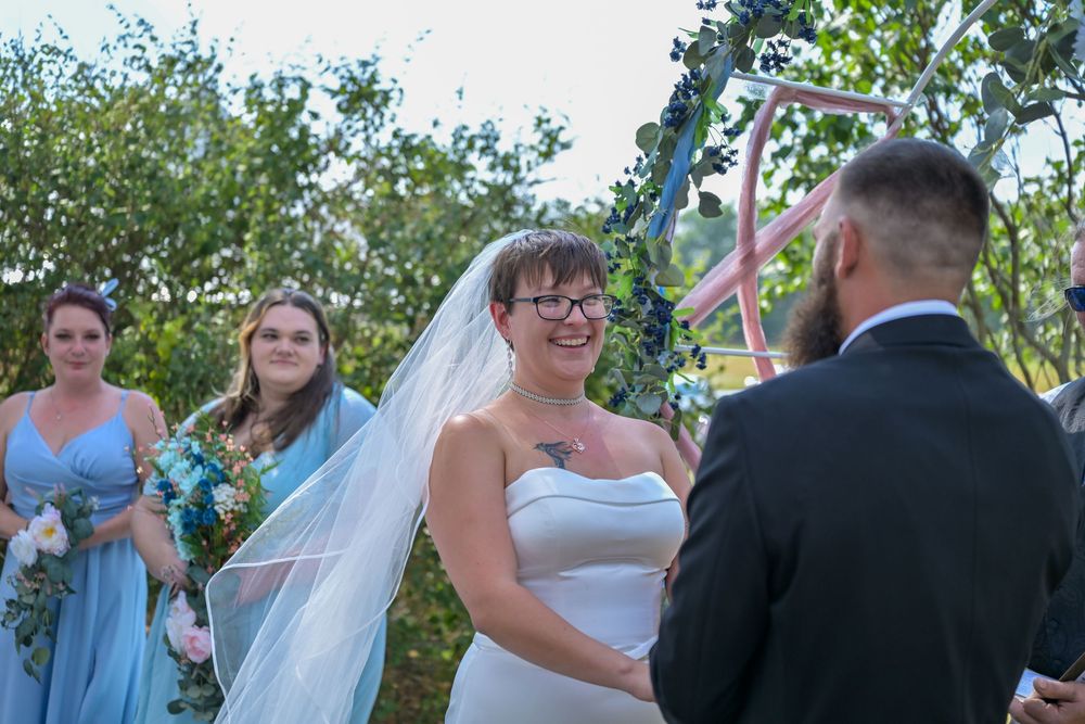 Bride in white gown and veil smiles at groom during outdoor wedding ceremony with bridesmaids in blue dresses.