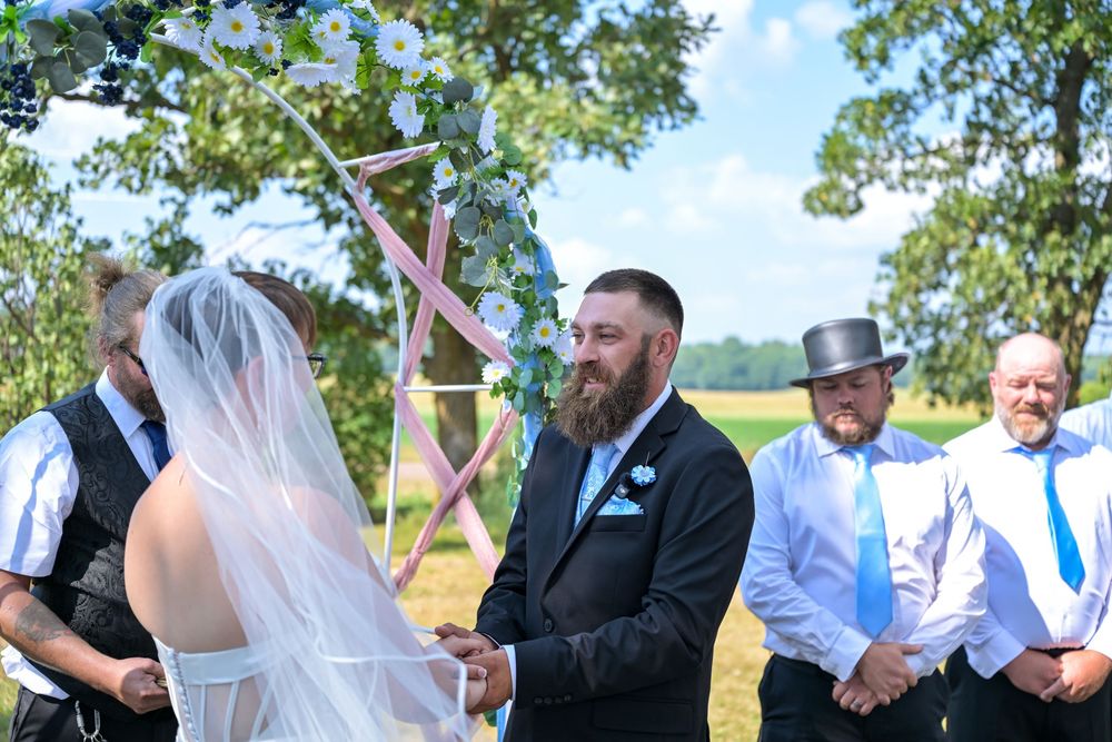Bearded groom in black suit smiles at bride during outdoor wedding ceremony with groomsmen and floral arch backdrop.