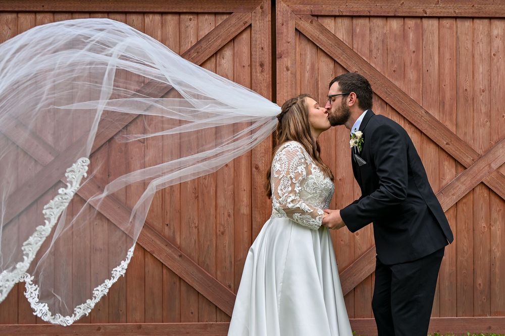 Bride and groom share a romantic kiss outdoors, with wedding veil blowing in wind against rustic barn doors.