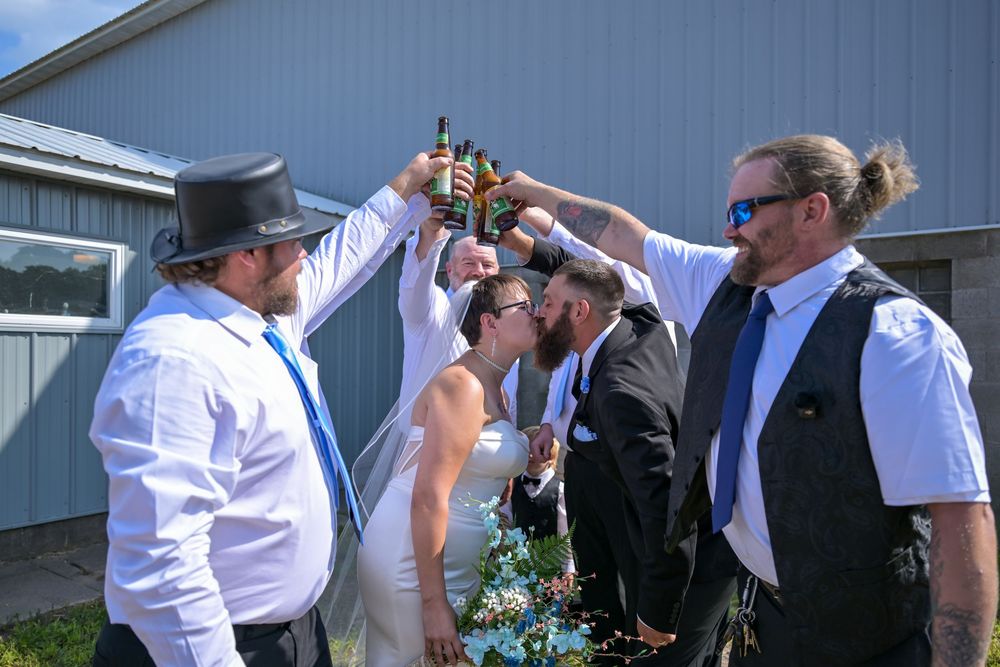 Wedding party toasting with beer bottles outdoors, bride and groom kissing while groomsmen celebrate in blue ties.