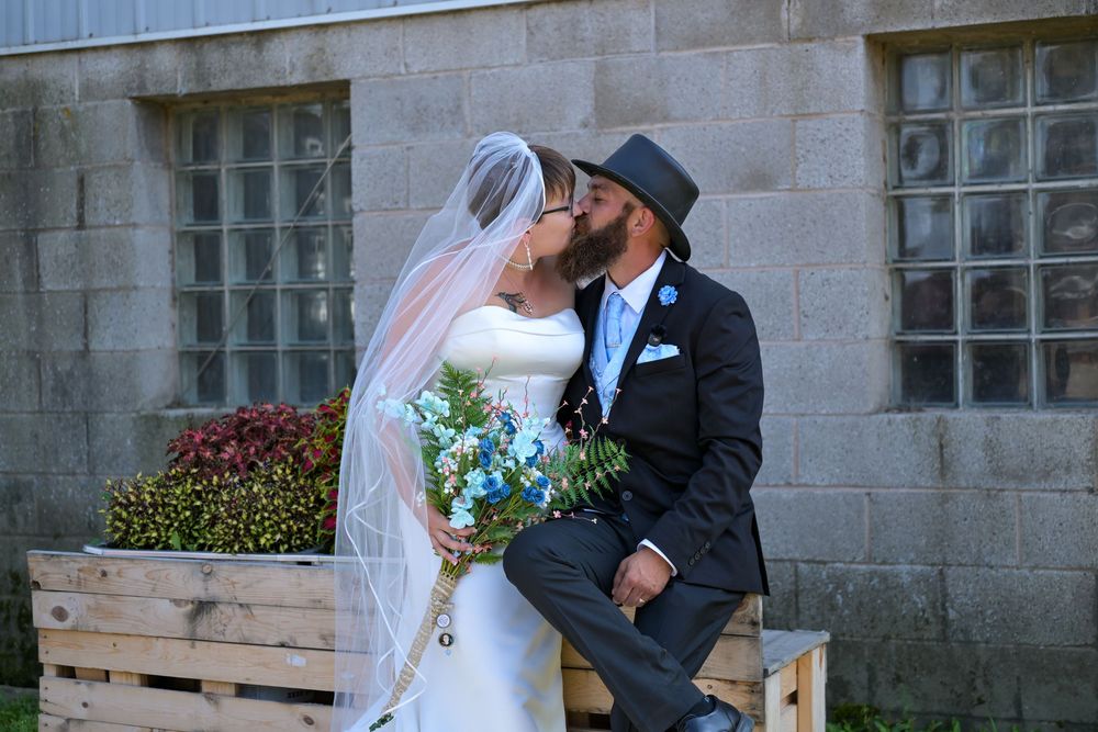 A bride and groom share a kiss outdoors, she holds blue flowers, he wears a gray hat and navy suit.