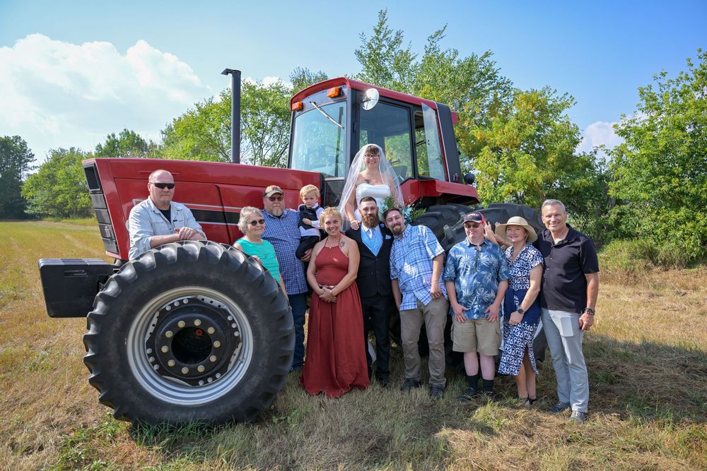 Wedding party poses with a red tractor in a green field on a sunny day.