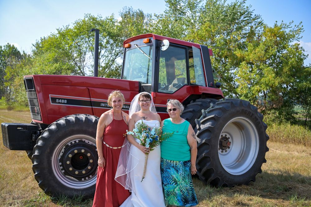Bride in white gown poses with two women in front of a red farm tractor outdoors on a sunny day.