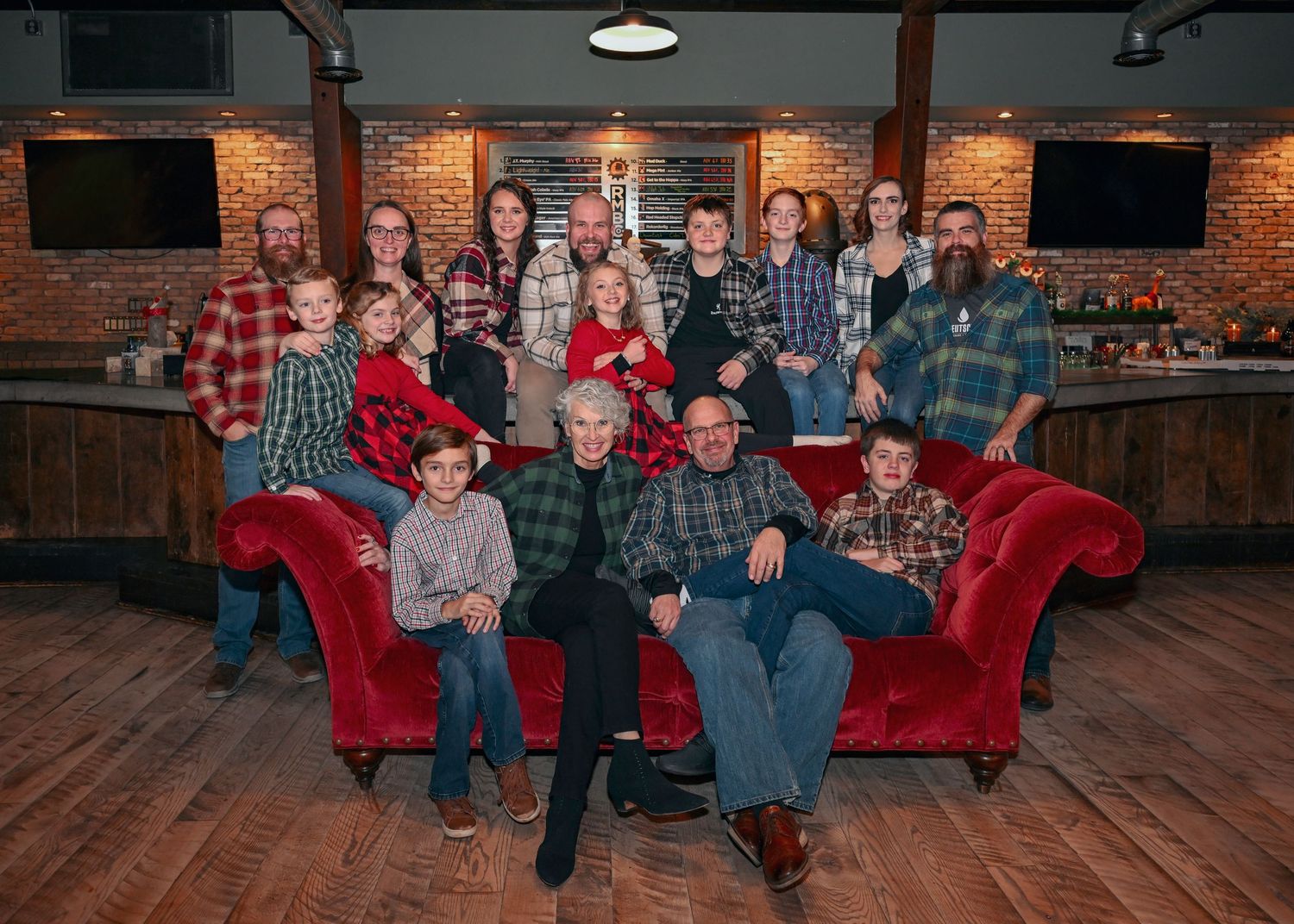 Large family group poses together on red velvet sofa in rustic bar venue wearing coordinated plaid outfits.