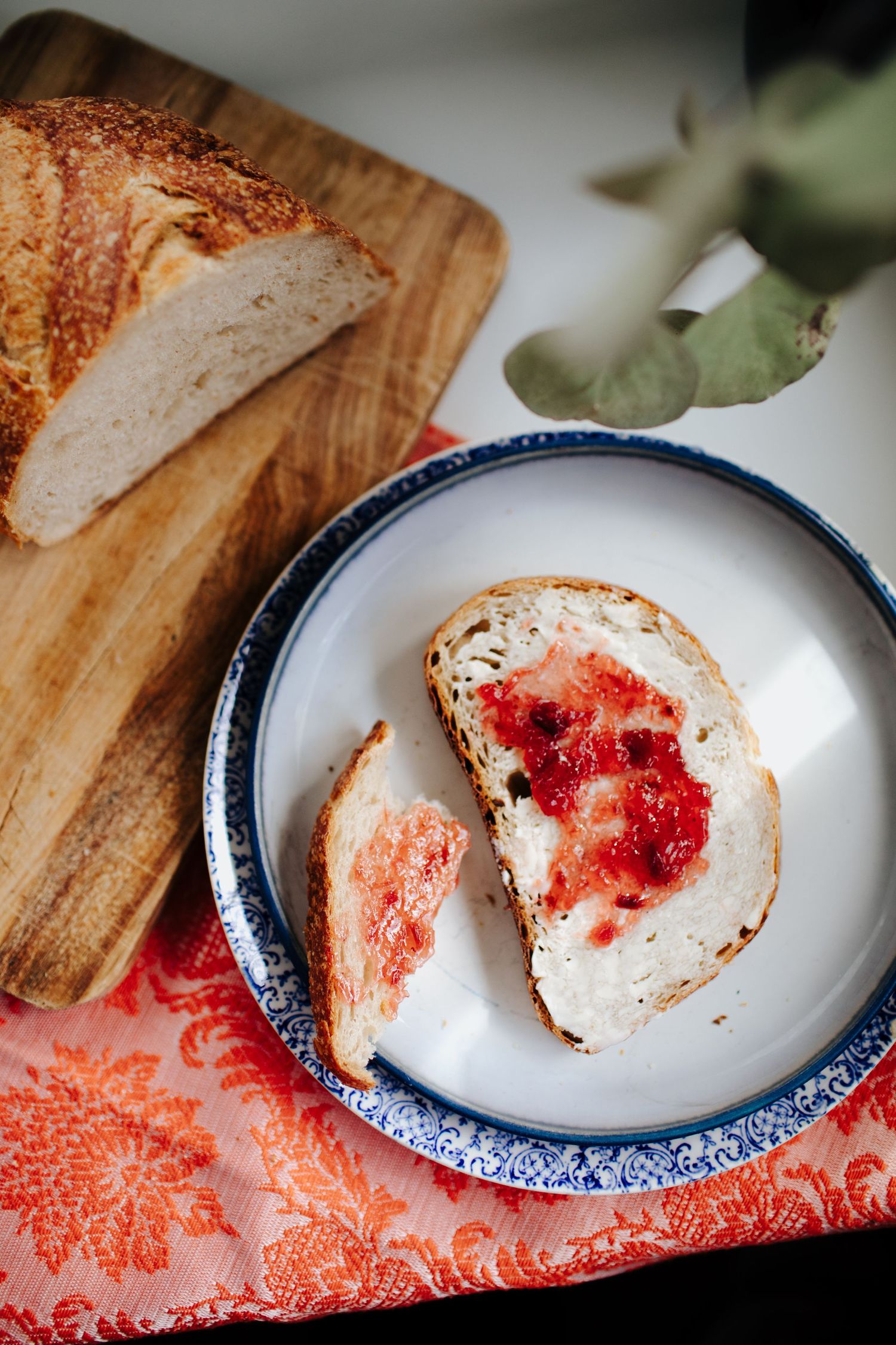 Sliced artisan bread with red jam spread on a blue-rimmed plate and wooden cutting board.