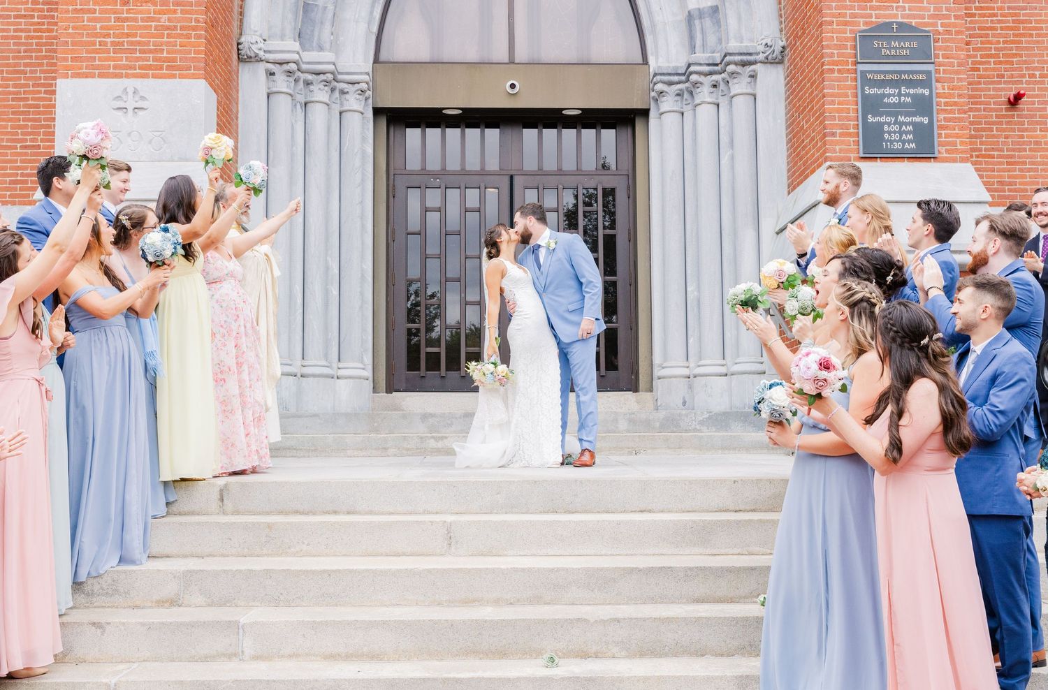Wedding party in pink and blue dresses creates celebratory pathway on church steps during exit.