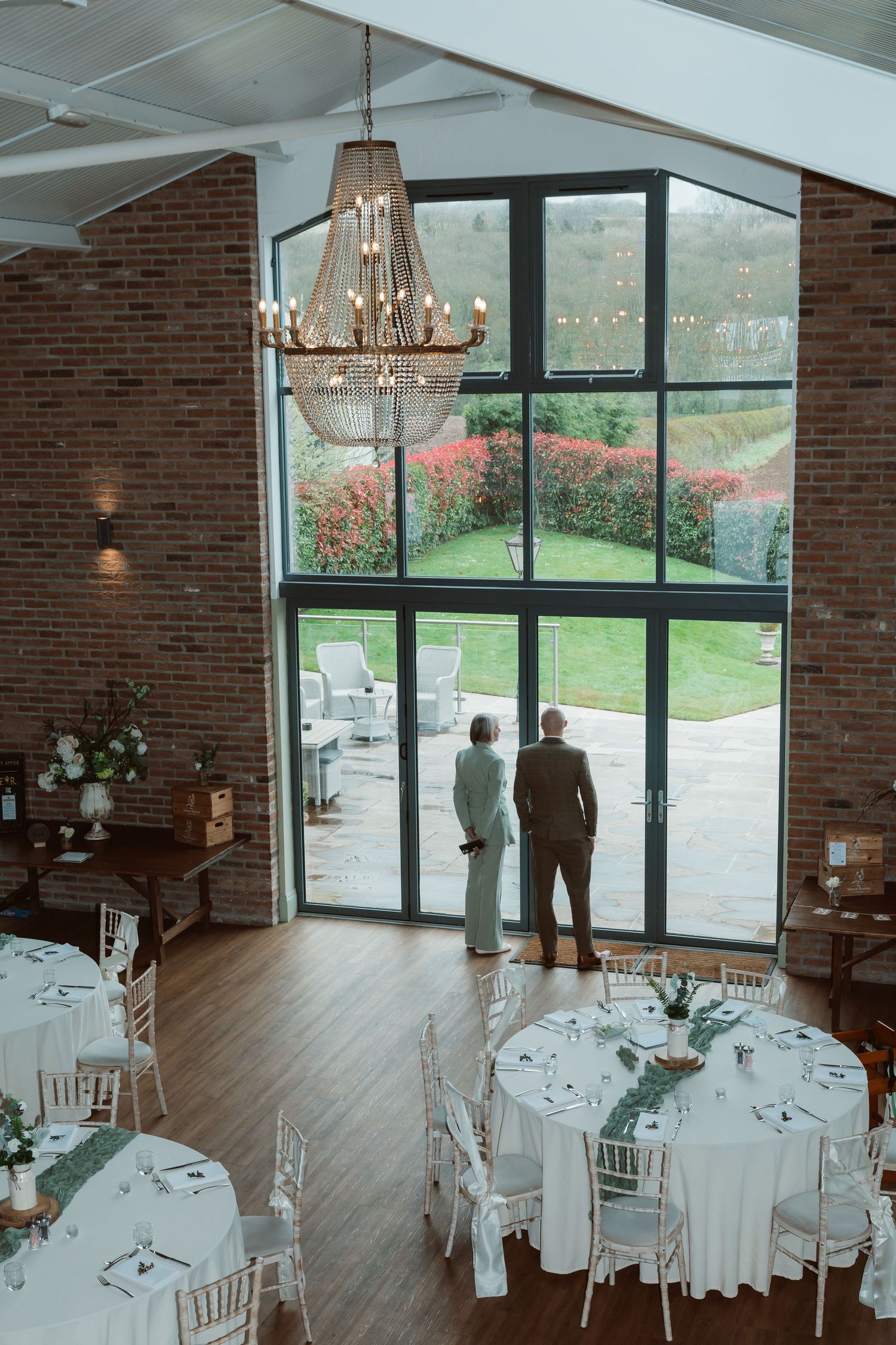 Elegant wedding reception venue with round tables, chandeliers, and floor-to-ceiling windows overlooking a garden. Photographer. Hull. East