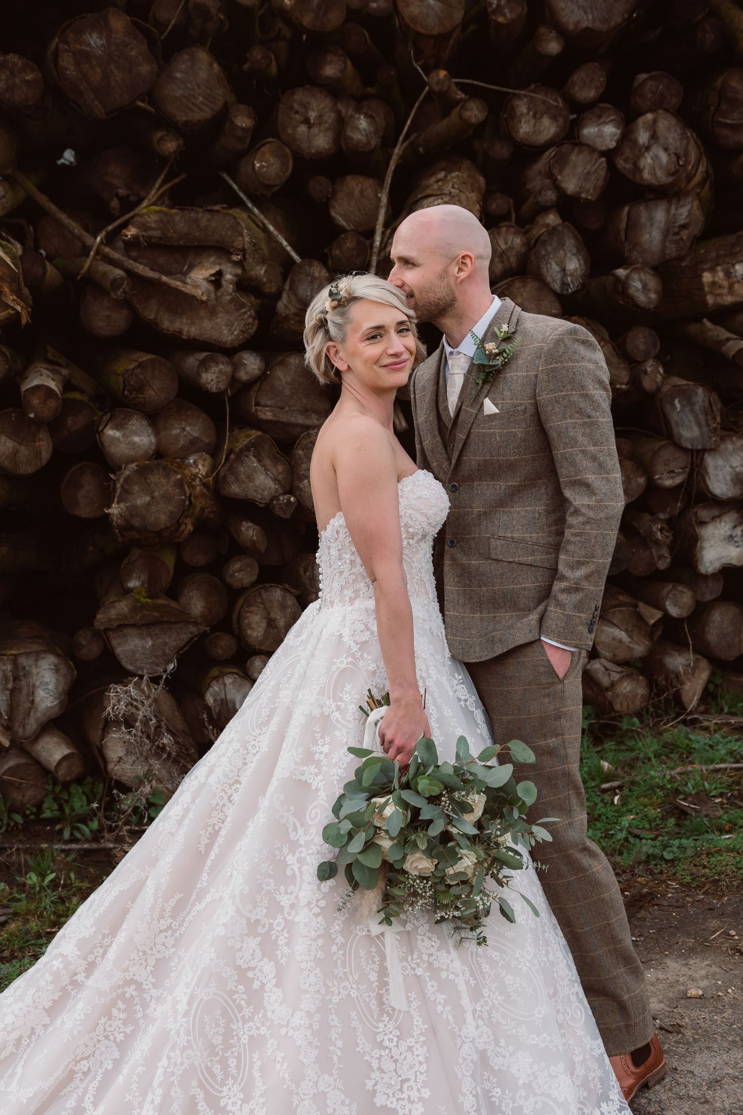 Newlyweds share an intimate moment against a rustic woodpile backdrop with eucalyptus bouquet. Photographer. Hull. East Yorkshire. Photo