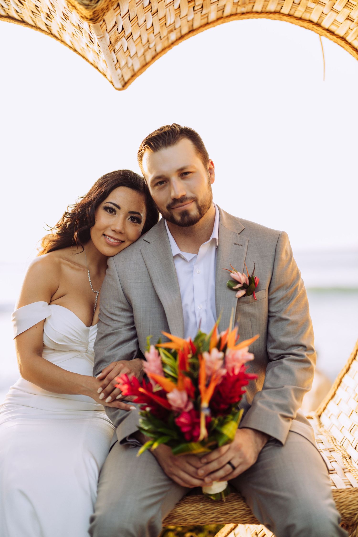 Wedding couple seated under woven arch with tropical flowers at beach ceremony.