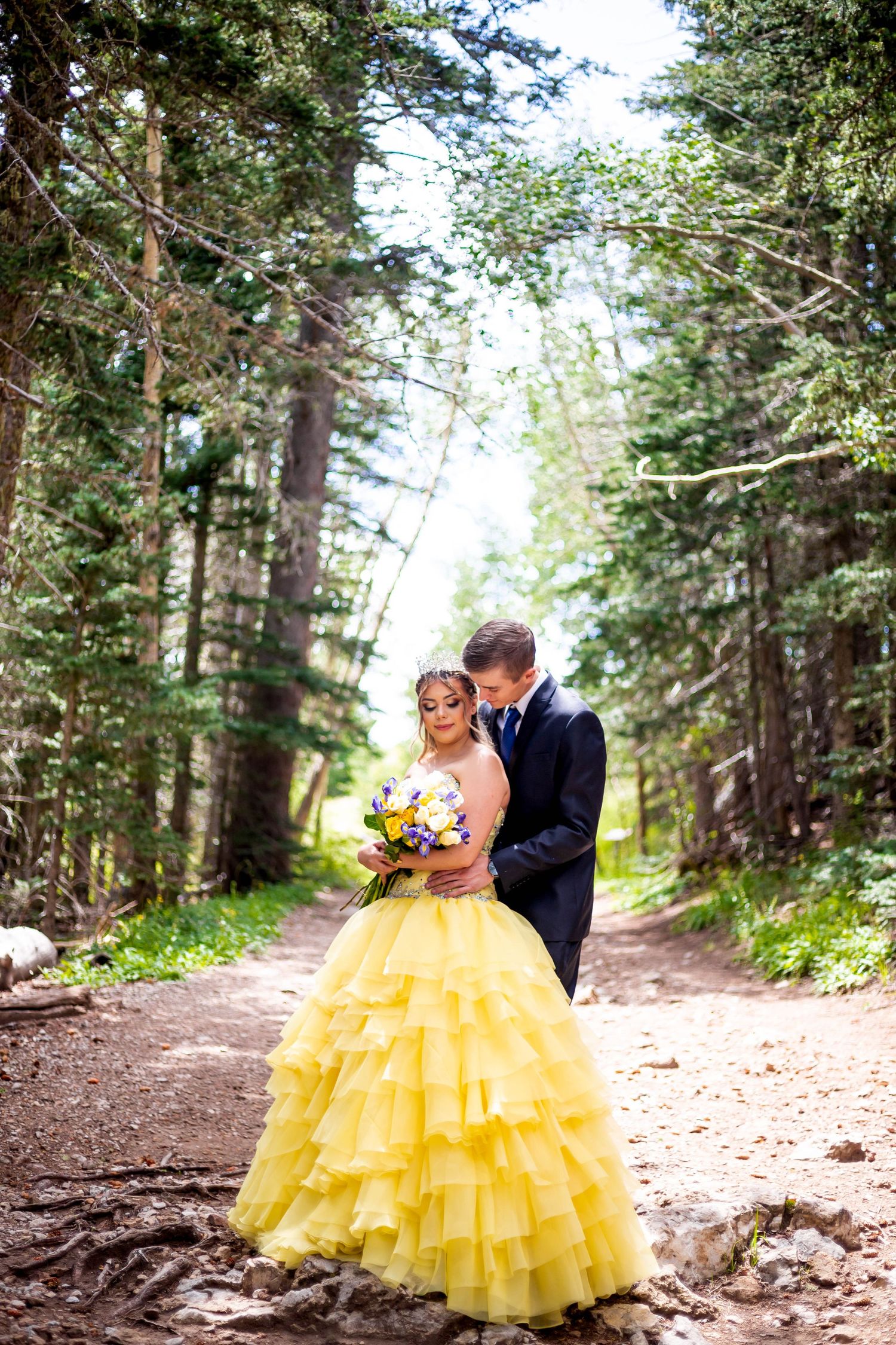 Bride in yellow ballgown and groom embrace on forest path during wedding photoshoot.