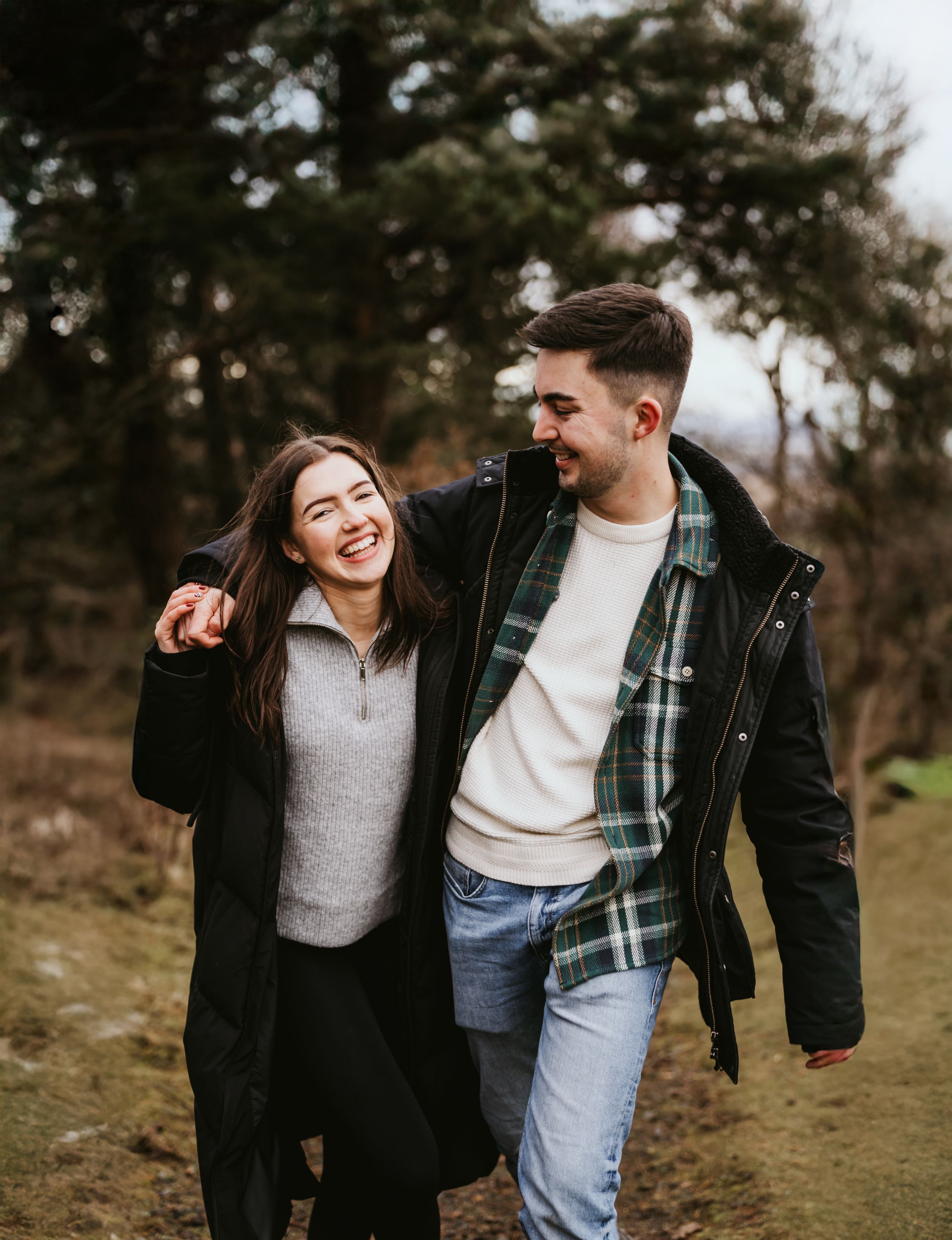 Happy couple embracing and laughing together while walking outdoors in a wooded area on an overcast day.