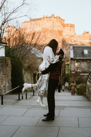 Romantic scene with couple embracing in front of Edinburgh Castle during golden sunset on cobblestone street.