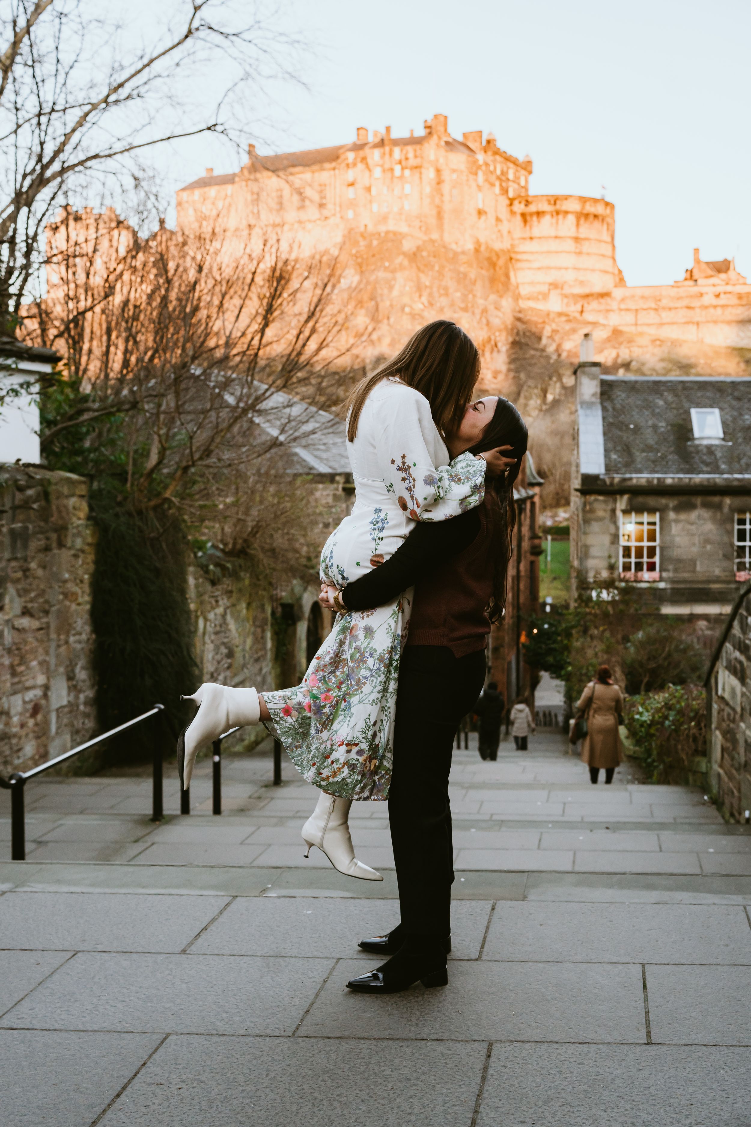 Romantic scene with couple embracing in front of Edinburgh Castle during golden sunset on cobblestone street.