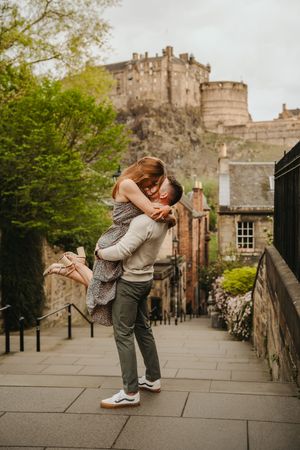 A romantic couple embraces near Edinburgh Castle on a stone walkway surrounded by historic architecture.