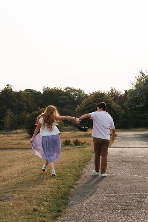 A couple walks joyfully down a dirt path together at golden hour while holding hands and leaning away from each other.