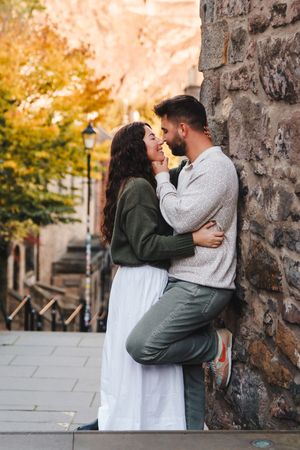 A romantic couple embraces against a stone wall on an autumn street with golden foliage in the background.