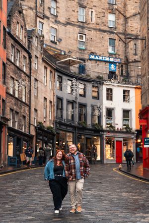 Historic cobblestone street in Edinburgh's Old Town with traditional stone buildings and storefronts on a cloudy day.