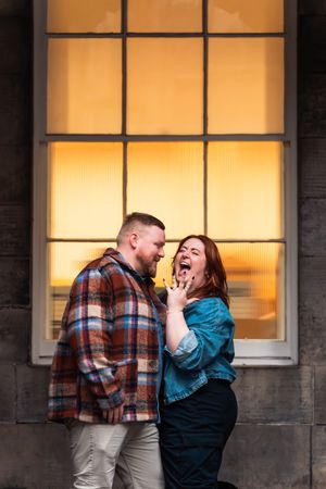 A couple shares a joyful moment together in front of illuminated windows at dusk.