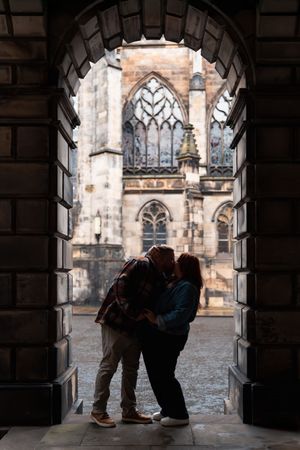 A couple shares a romantic kiss under a stone archway with a gothic cathedral visible in the background.