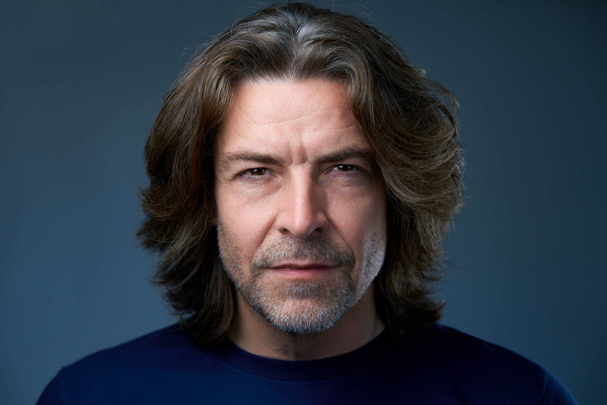 Person with dark tousled hair wearing dark clothing against blue background in portrait series.