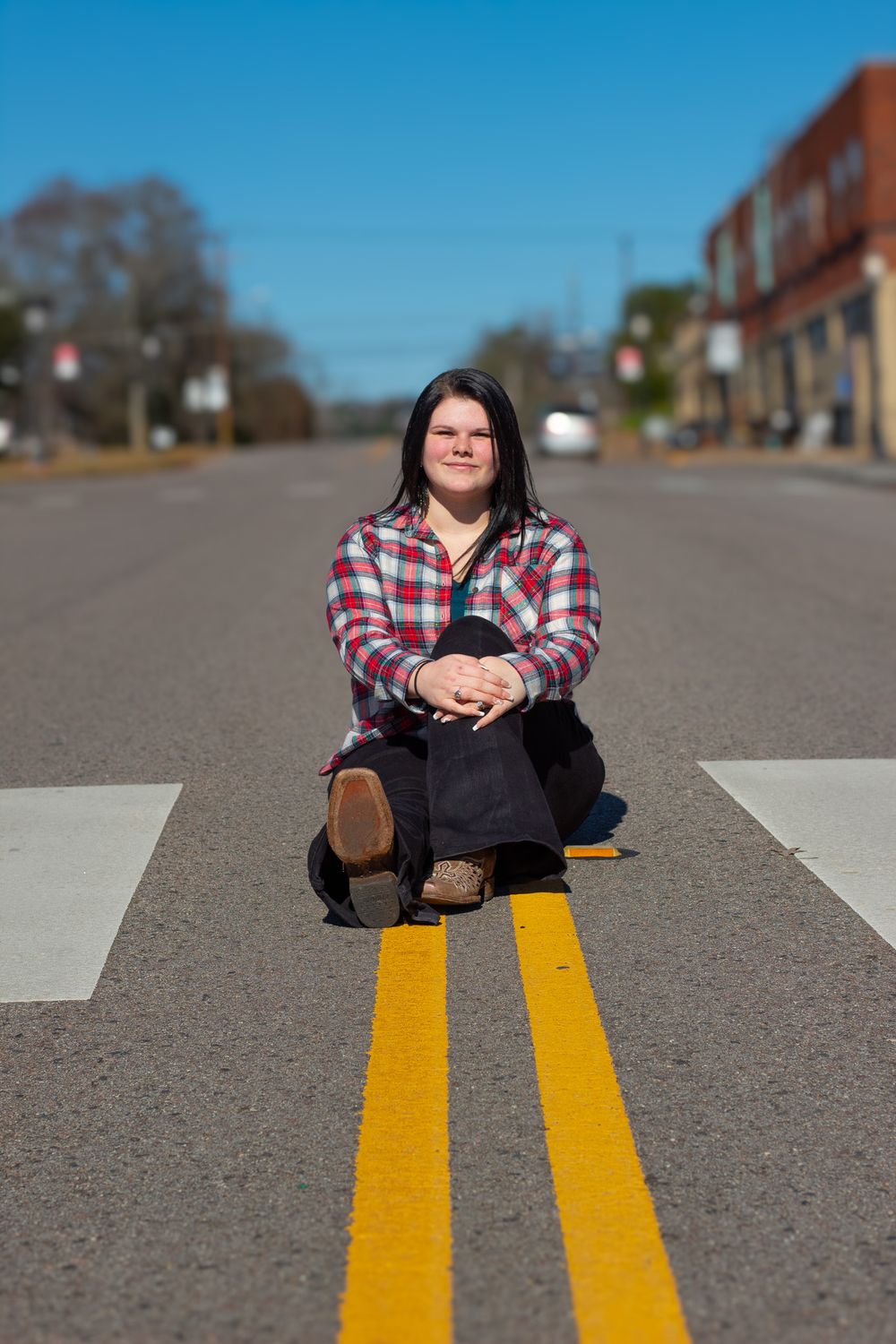 Someone sits on yellow road lines in the middle of a small town street wearing a red plaid flannel shirt and dark pants.